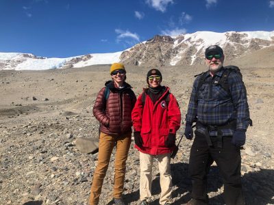 3 people stand on rocky, snowy mountain plateau dressed in cold weather attire